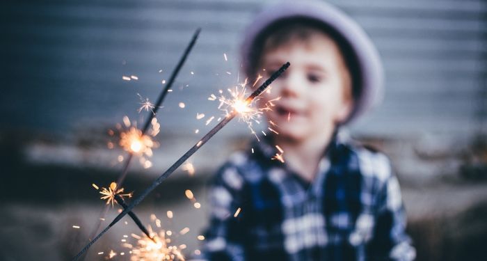 a small child holding a sparkler. The sparkler is clear in the foreground; the boy, who is wearing a grey hat and a blue plaid button-up shirt, is blurred in the background