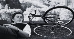 a white man in a white button down and a black vest laying in the grass net to a bicycle, holding a book in his hands