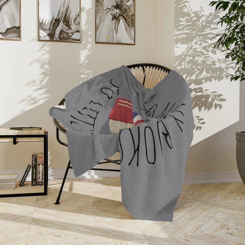 Photo of a grey blanket open on top of a chair. The blanket has an illustration of a couple of colourful books and the text So many books so little time in black letters all across it