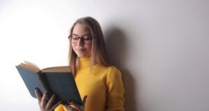 white woman with yellow sweater reading a book against a white wall