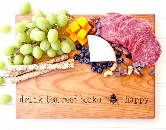 Wooden cutting board topped with cheese and meats, etched with phrase "drink tea, read books, bee happy"