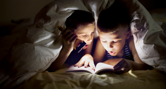 a photo of two kids reading under covers with a flashlight, looking shocked at the book