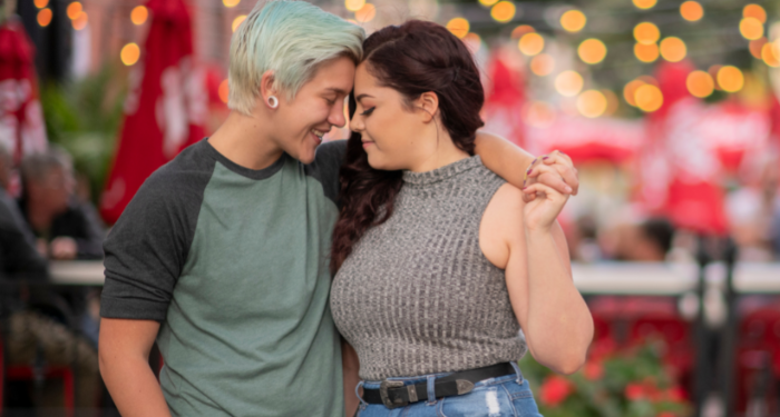 a photo of a sapphic couple holding hands, leaning their foreheads together, and smiling, with lights in the background