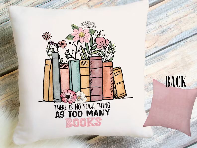 Photo of a pillow with a colourful print of books standing side by side, showing pink, yellow and blue spines, a few pink and white flowers, and below the text there is no such thing as too many books. 