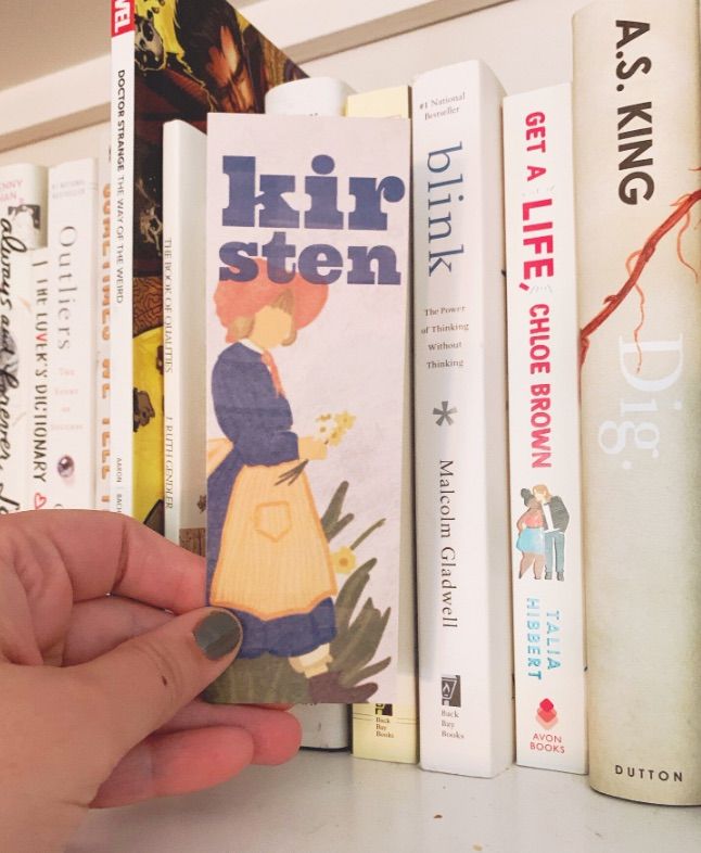 Image of a bookmark with an illustration of Kiersten. It is being held by a white hand in front of a shelf of white books. 