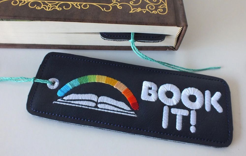 Image of a black bookmark. It is embroidered with the book it logo. 