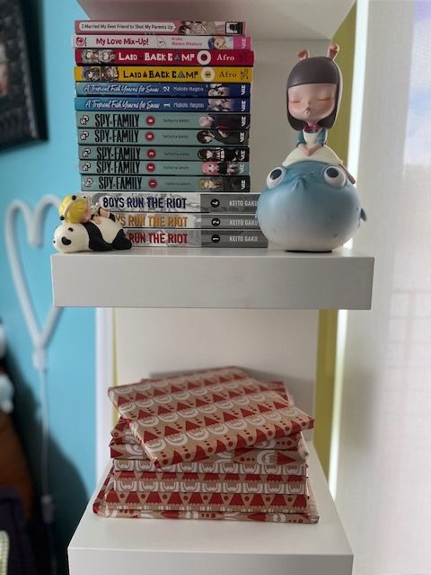 bookshelf with a stack of manga, art toys, and a stack of graphic novels wrapped in Santa Clause wrapping paper.