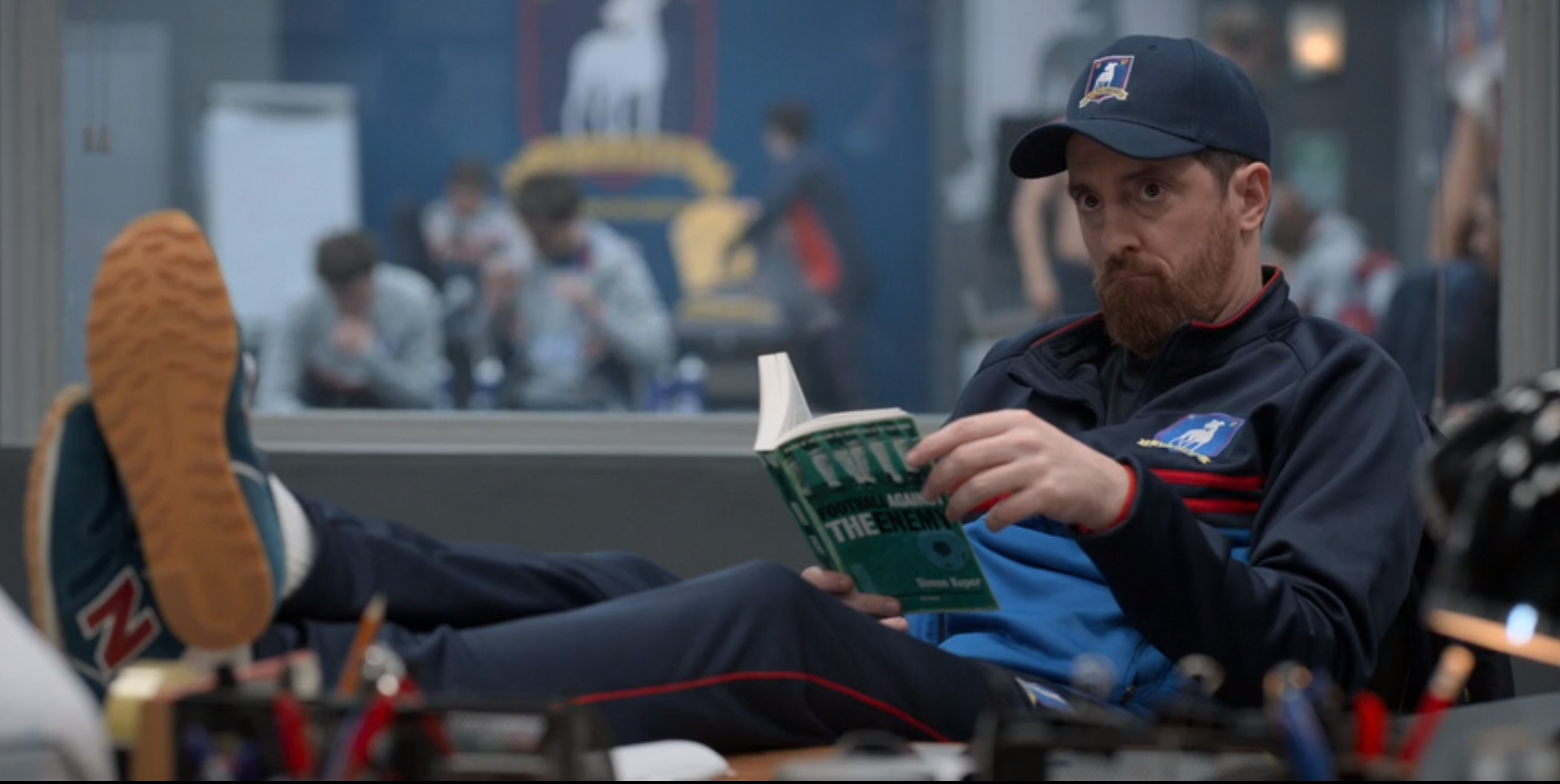 a still of Coach Beard reading a book with his feet up on the desk. He's looking up from the book at Ted.