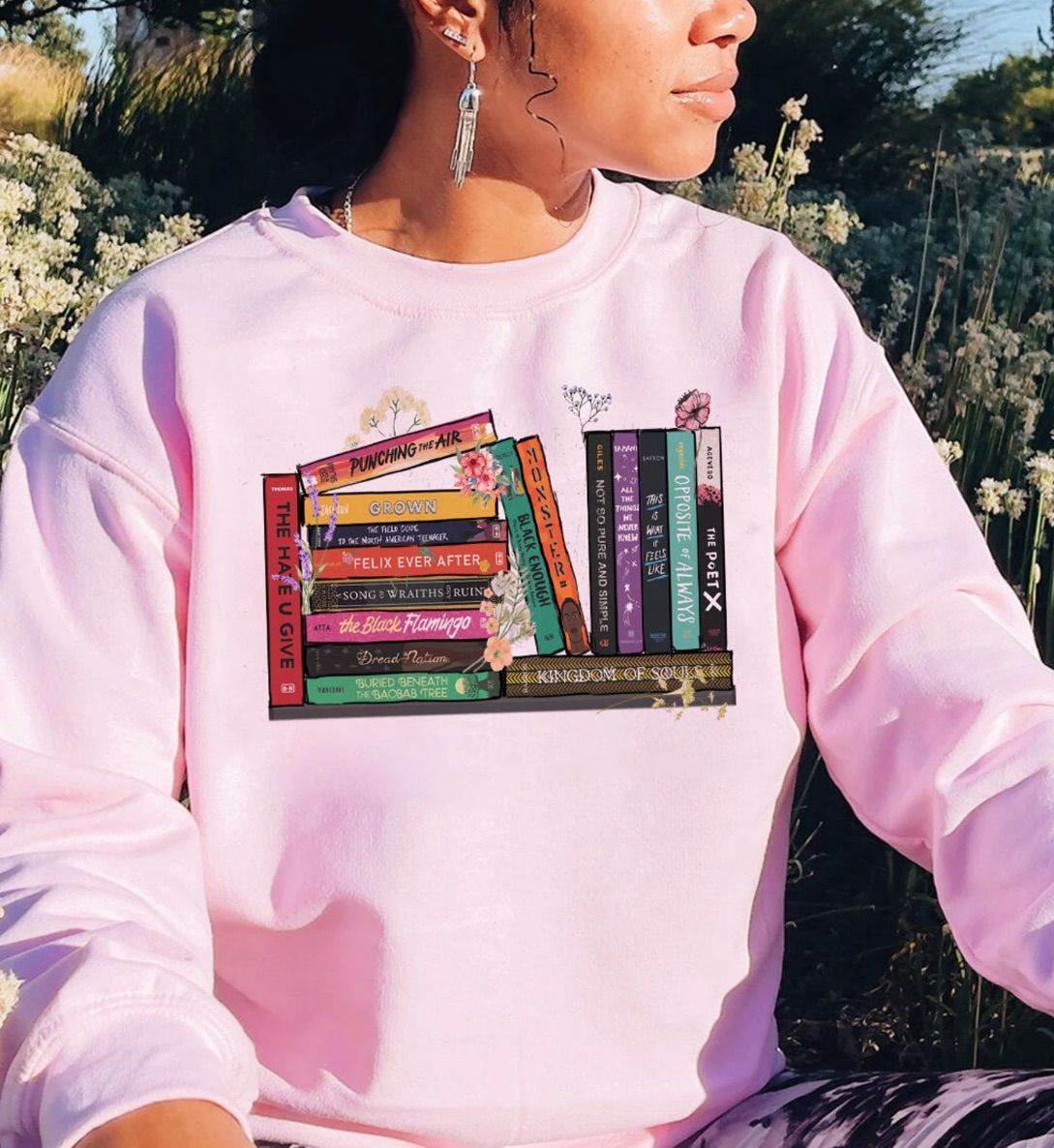 Image of a Black woman wearing a pink sweatshirt. The shirt has several Black YA book spines on it. 
