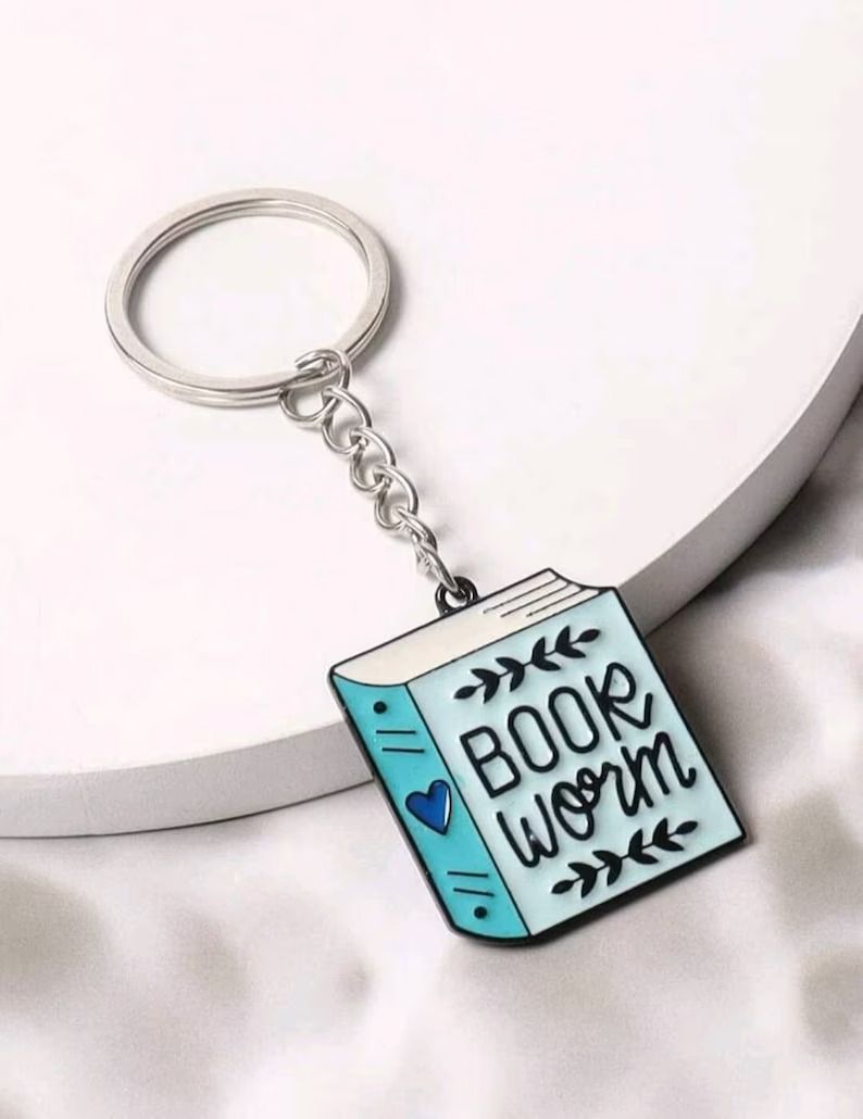 Photo of a keychain made of enamel in the shape of a book painted in blue, the words bookworm on the front.