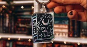 Photo of a black keychain in the shape of a book with silver drawings on the cover and spine. The cover has plants and the moon, the spine has the text Fairy Smut on it.