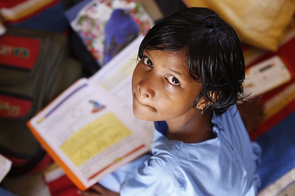 A child with brown skin and black hair, wearing a blue top, looks up at the camera. A book is open on their lap.