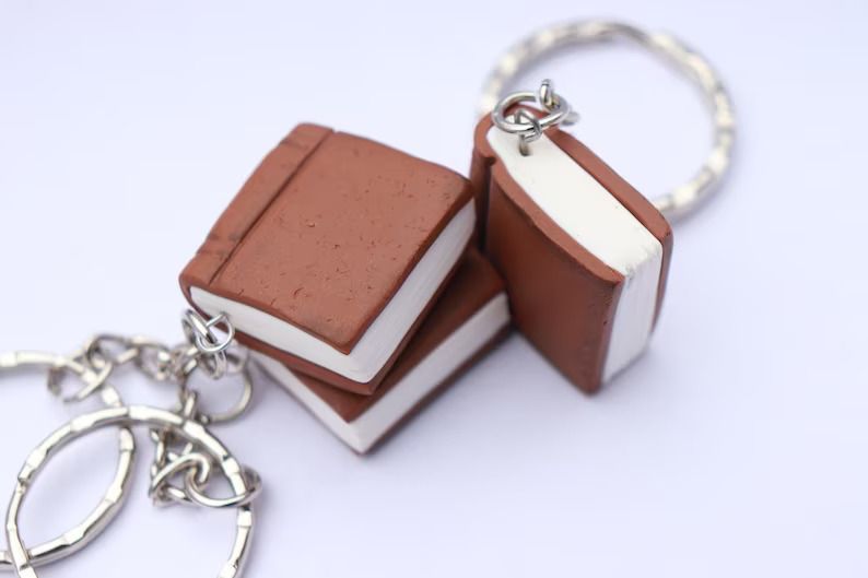 Photo of three small polymer clay keychains shaped like a book, imitating a leather bound book. 