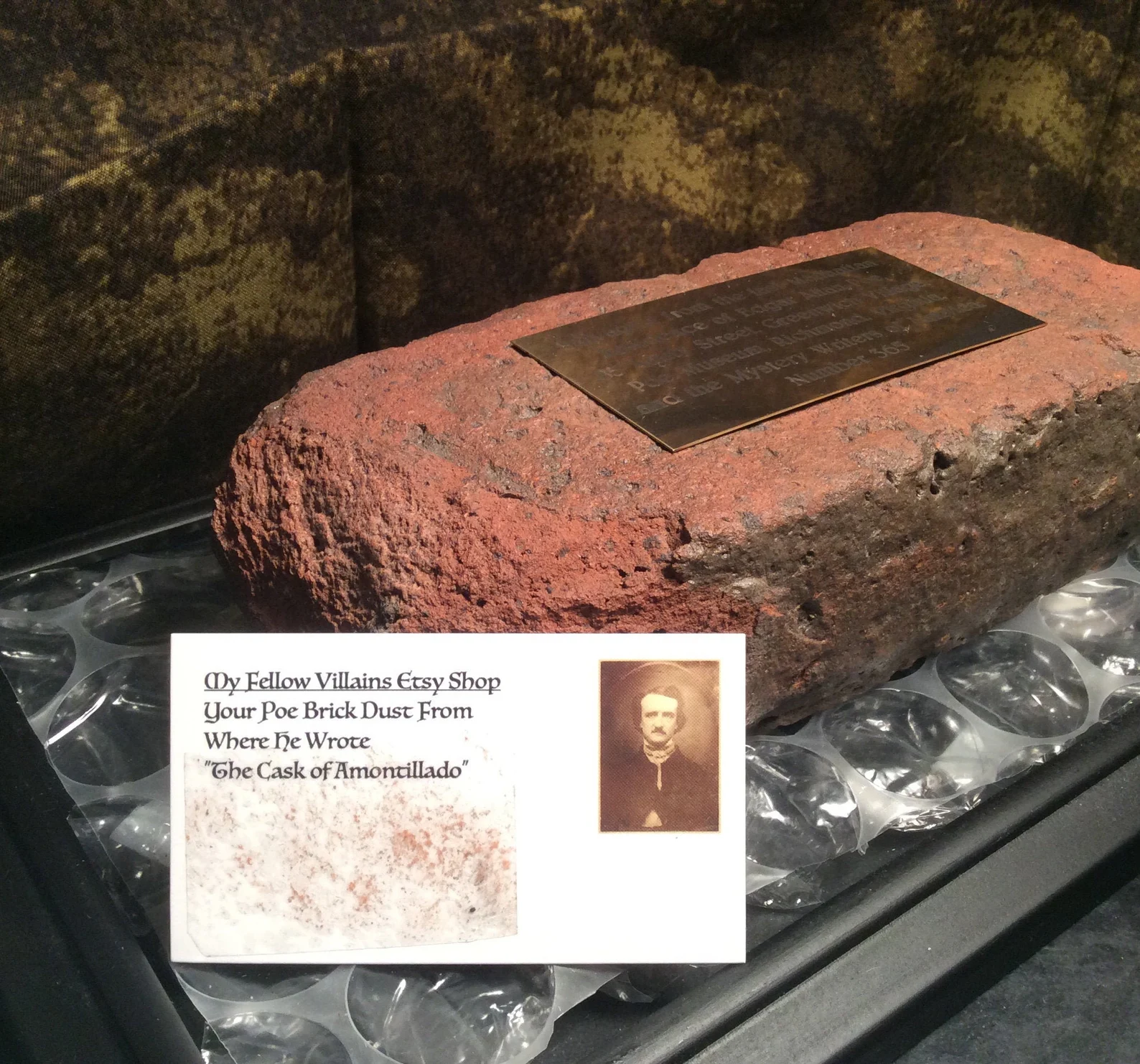 A brick with a brass plaque on it declaring the brick to be from the place Poe wrote The Cask of Amontillado, and a business card with dust from the brick affixed to it.