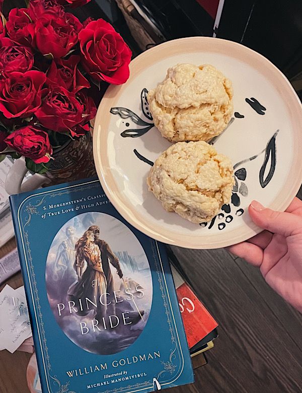 A photo of a hand holding a plate with two scones on it, held over The Princess Bride and beside red roses