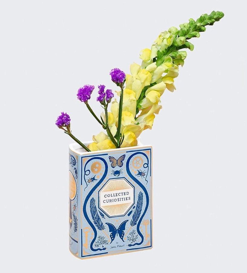 Image of several flowers inside a vase in the shape of a book. The book is blue and gold, and it is titled "collected curiosities."