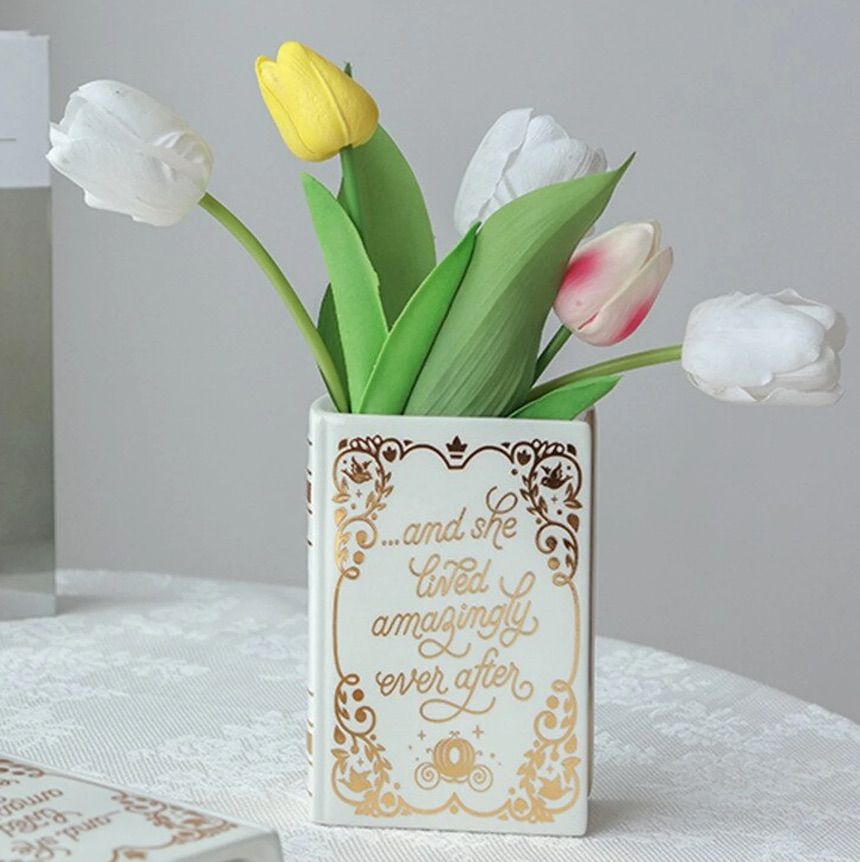 Image of a white vase holding tulips. The vase in the shape of a book has gold text that reads "and she lived amazingly ever after."