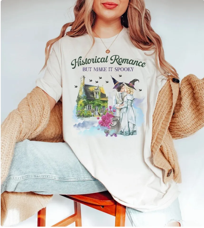 Image of a t-shirt featuring spooky horror--there are two witches kissing in front of a haunted house. 