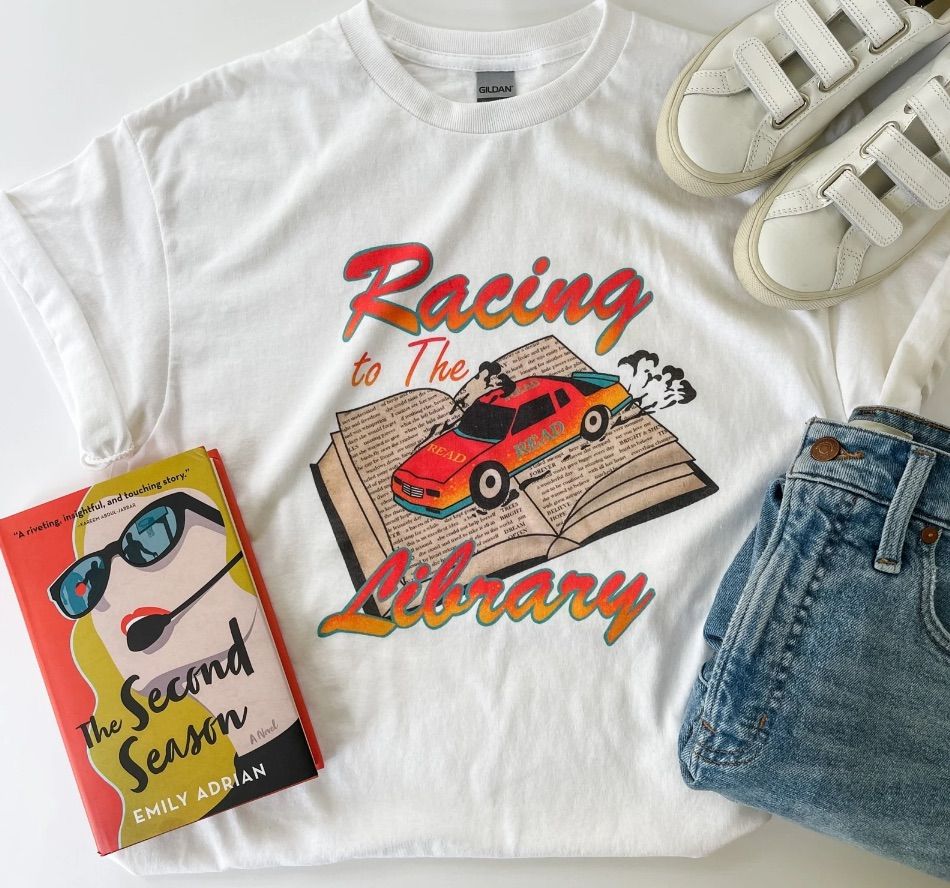 Image of a white t-shirt. It has a car driving on top of an open book with the words "racing to the library."