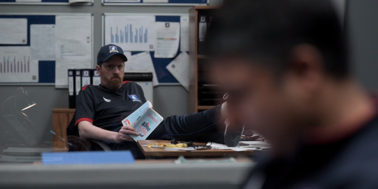 a still of Beard sitting with feet on his desk, holding About a Boy, and looking at Nate in the foreground with worry