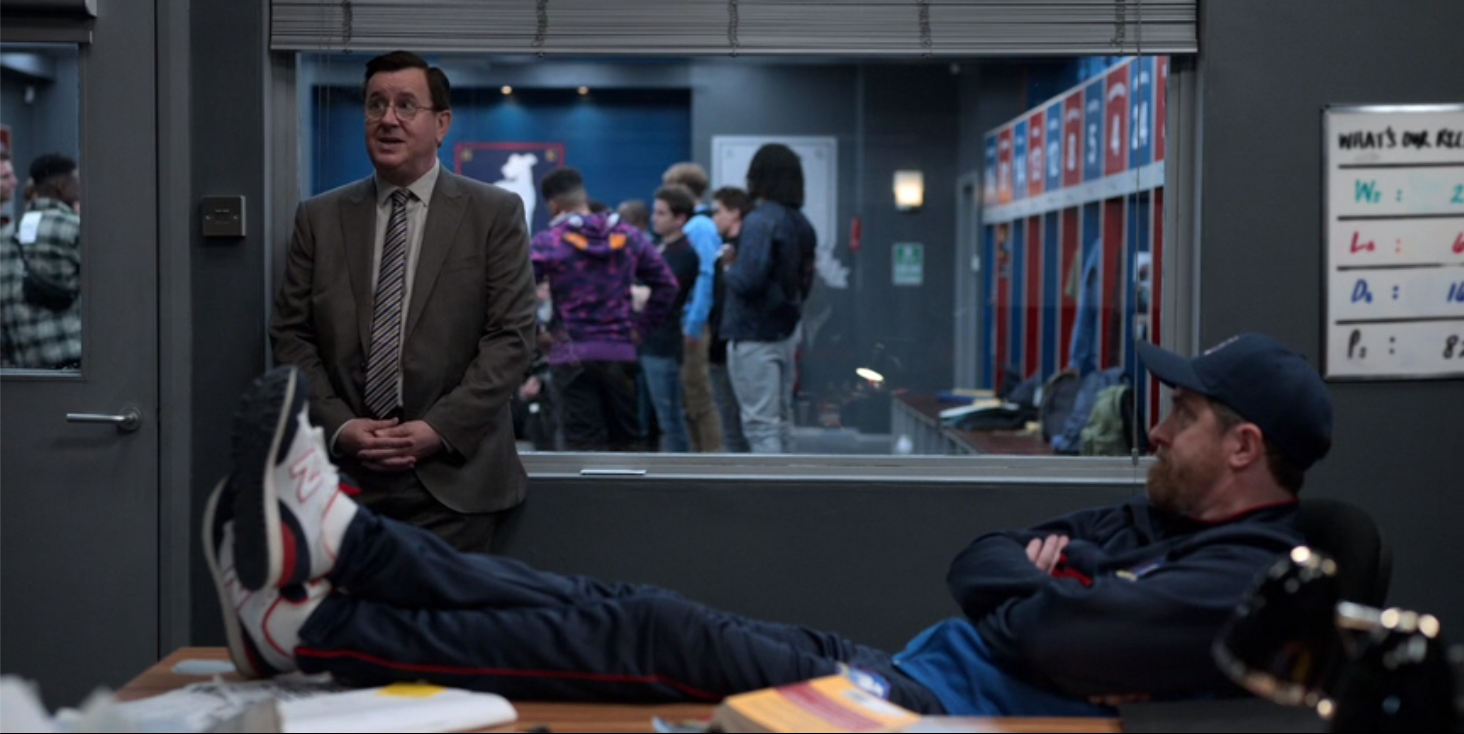 a still from Ted Lasso. Beard has his feet on his desk. Leslie is talking. The team is visible through the window, having a locker room chat. A book is facedown on Beard's desk.