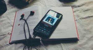 an open hardcover book with a phone connected to ear buds resting on top. The phone display is of a song in a music streaming app