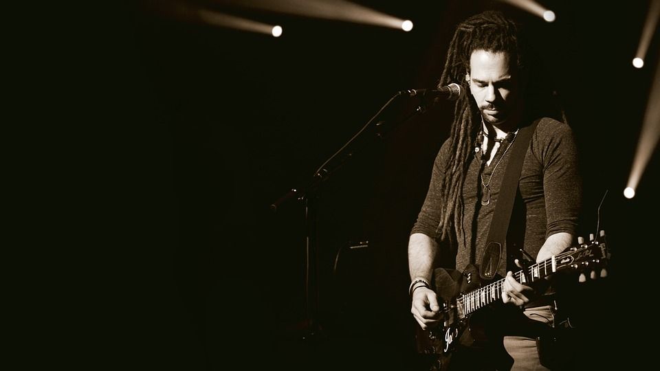 A black-and-white photo of a man with long hair in locks and a mid-long-sleeved shirt, playing a guitar in front of a mic stand.