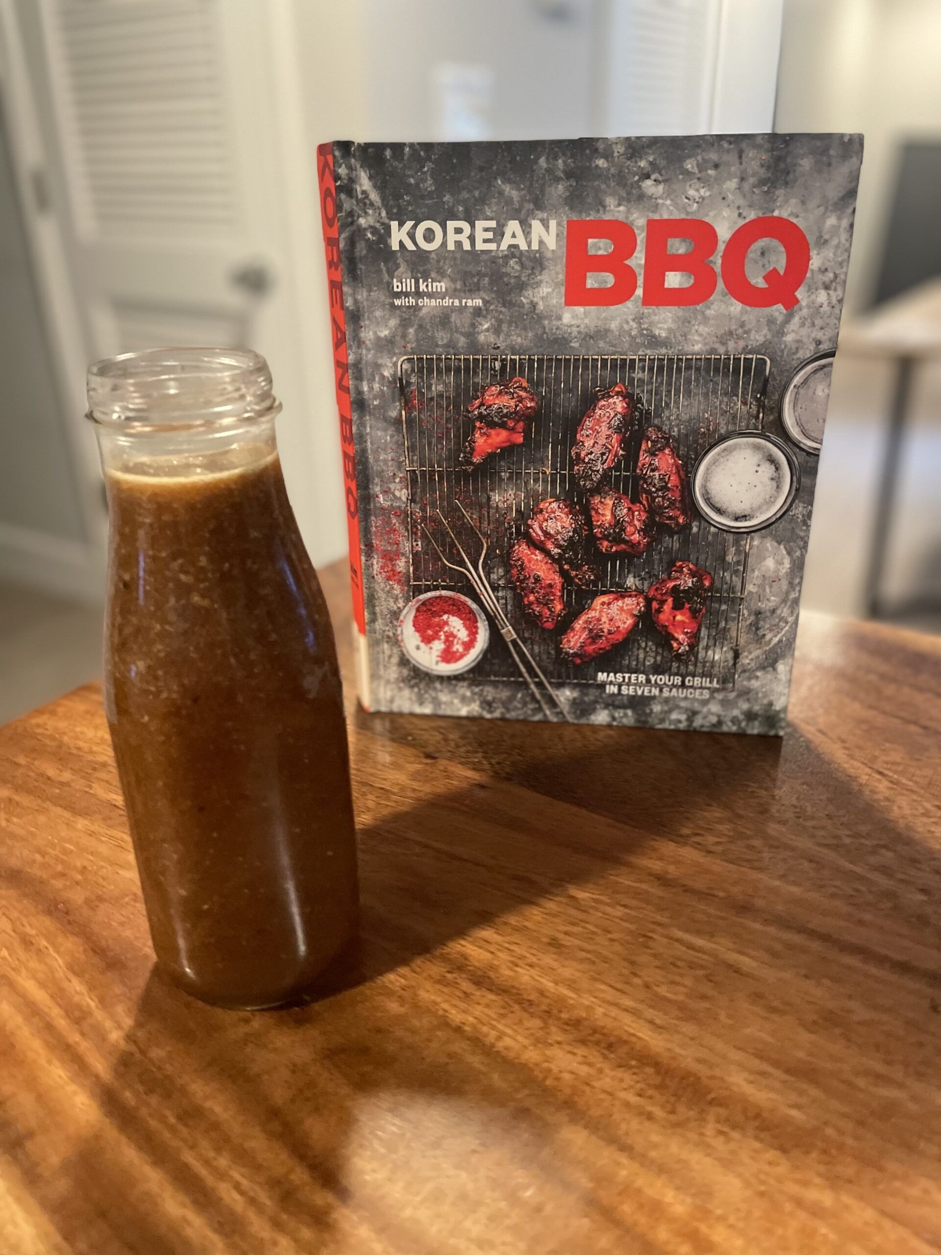 Image of Korean BBQ cookbook next to a tall glass jar of a light brownish BBQ sauce