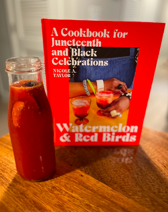 Photo of the cookbook Watermelon and Red Birds next to a tall glass jar of a thin reddish liquid with floating pieces of fig