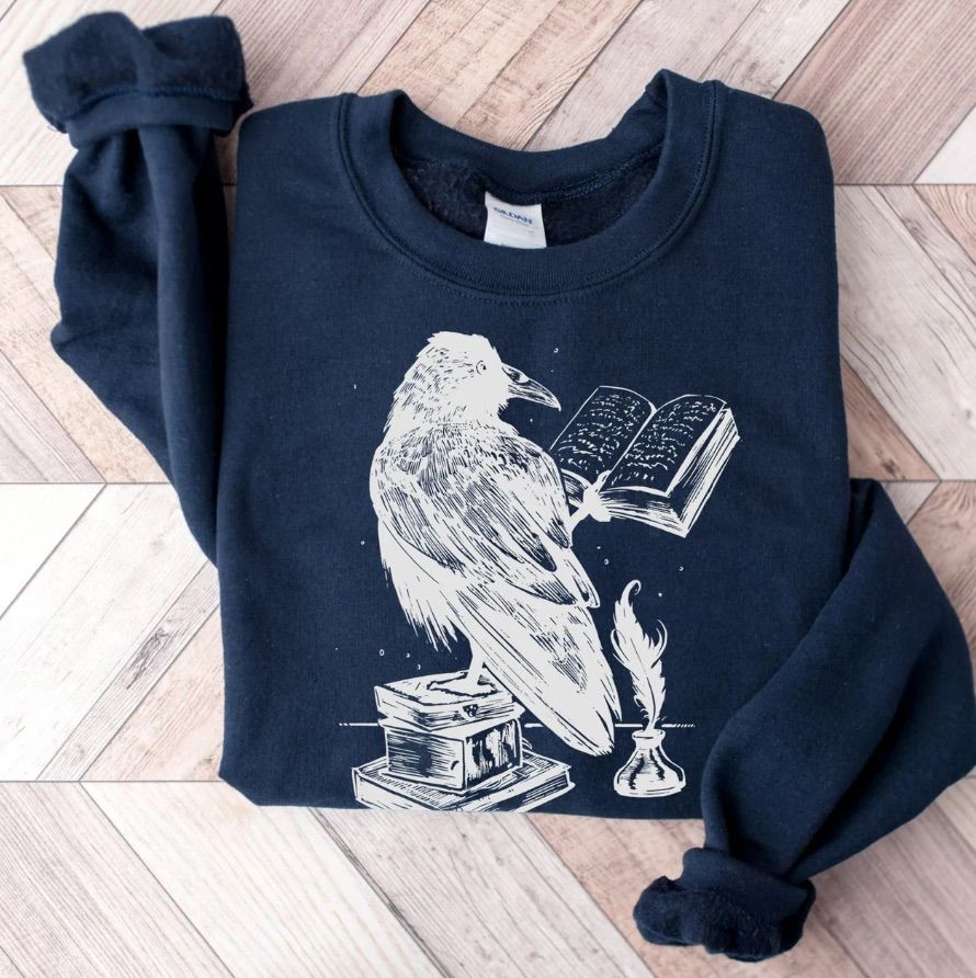 Image of a blue sweatshirt featuring a raven reading a book in white screen print. 