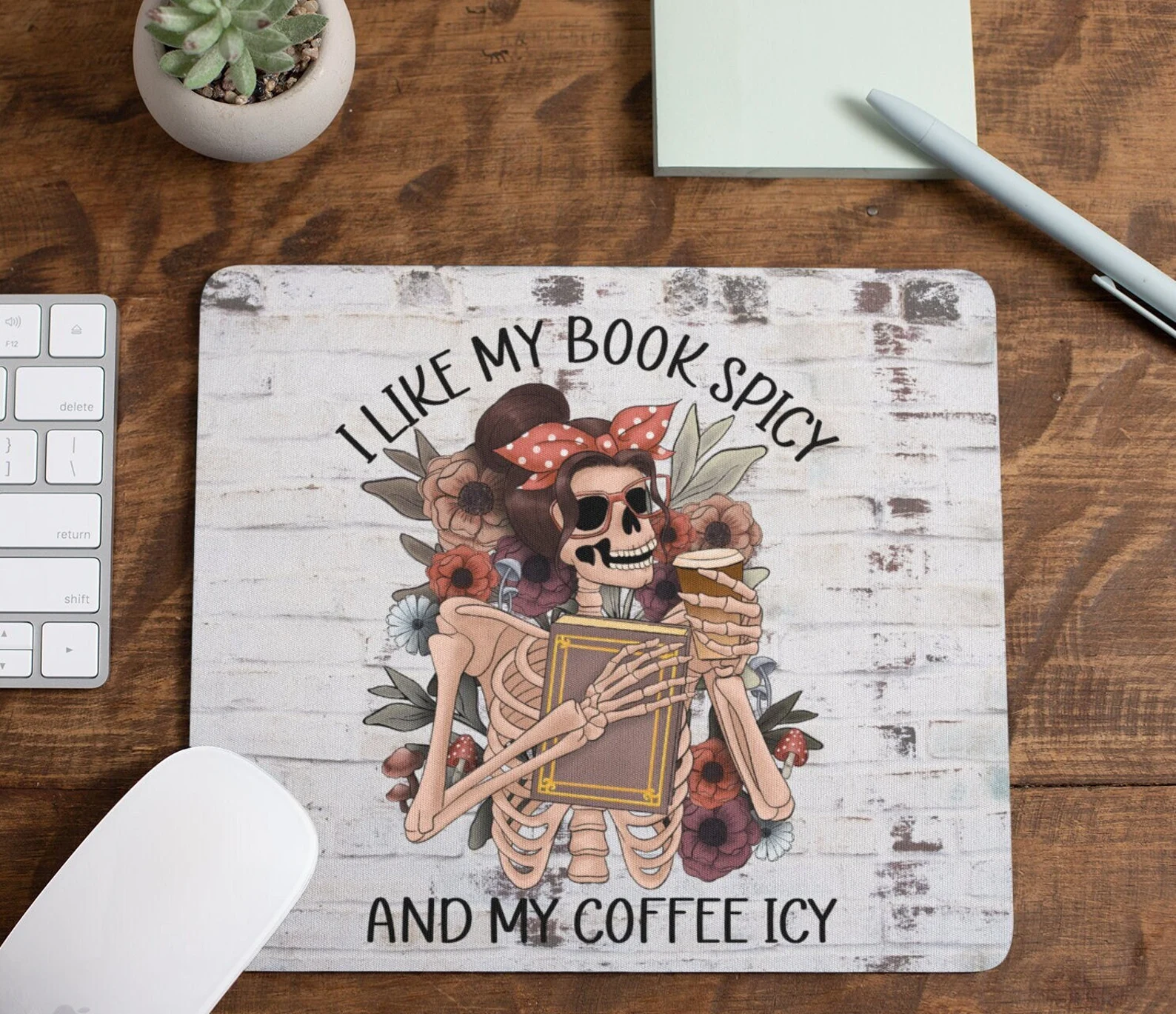 a mousepad with a skeleton with hair against a backdrop of flowers holding a book and iced coffee that reads "I like my books spicy and my coffee icy"