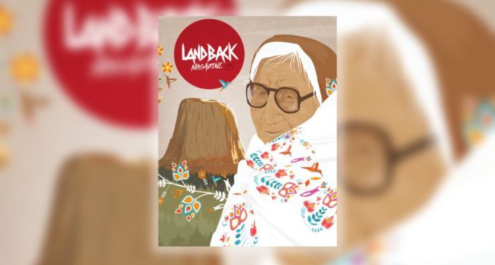 cover of the first edition of LANDBACK Magazine, showing an Indigenous person cloaked in a white garment with colorful embroidery with a mountain and grass in the background