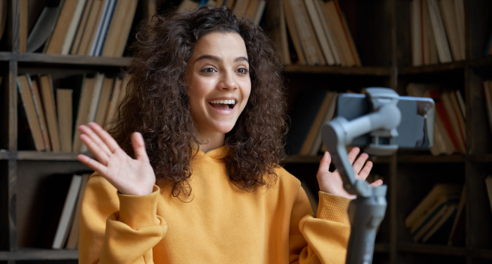 a picture of a woman in front of a bookshelf smiling and waving at a phone on a tripod