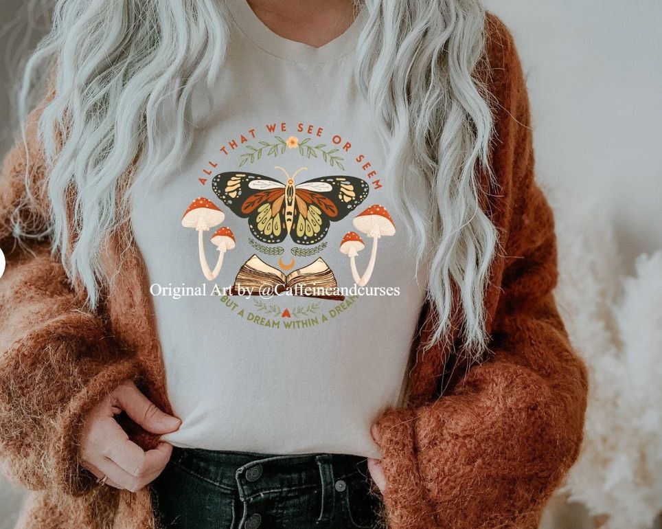 Image of a t-shirt featuring a book, butterfly, and several mushrooms. It has the text "all that we see or seem is but a dream within a dream."