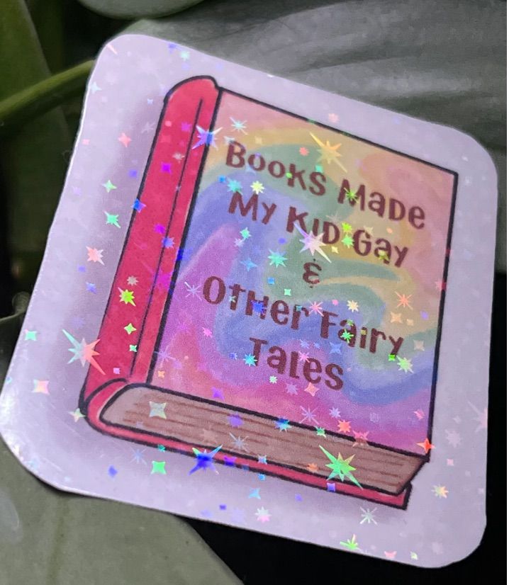 Image of a holographic sticker in the shape of a book that says "books made my kid gay and other fairy tales."