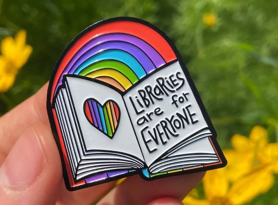 Image of an enamel pin in the shape of an open book that reads "libraries are for everyone."