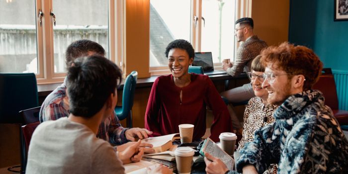 people of various skin tones gathered around a coffee shop table iwht drink cups and books, laughing with each other