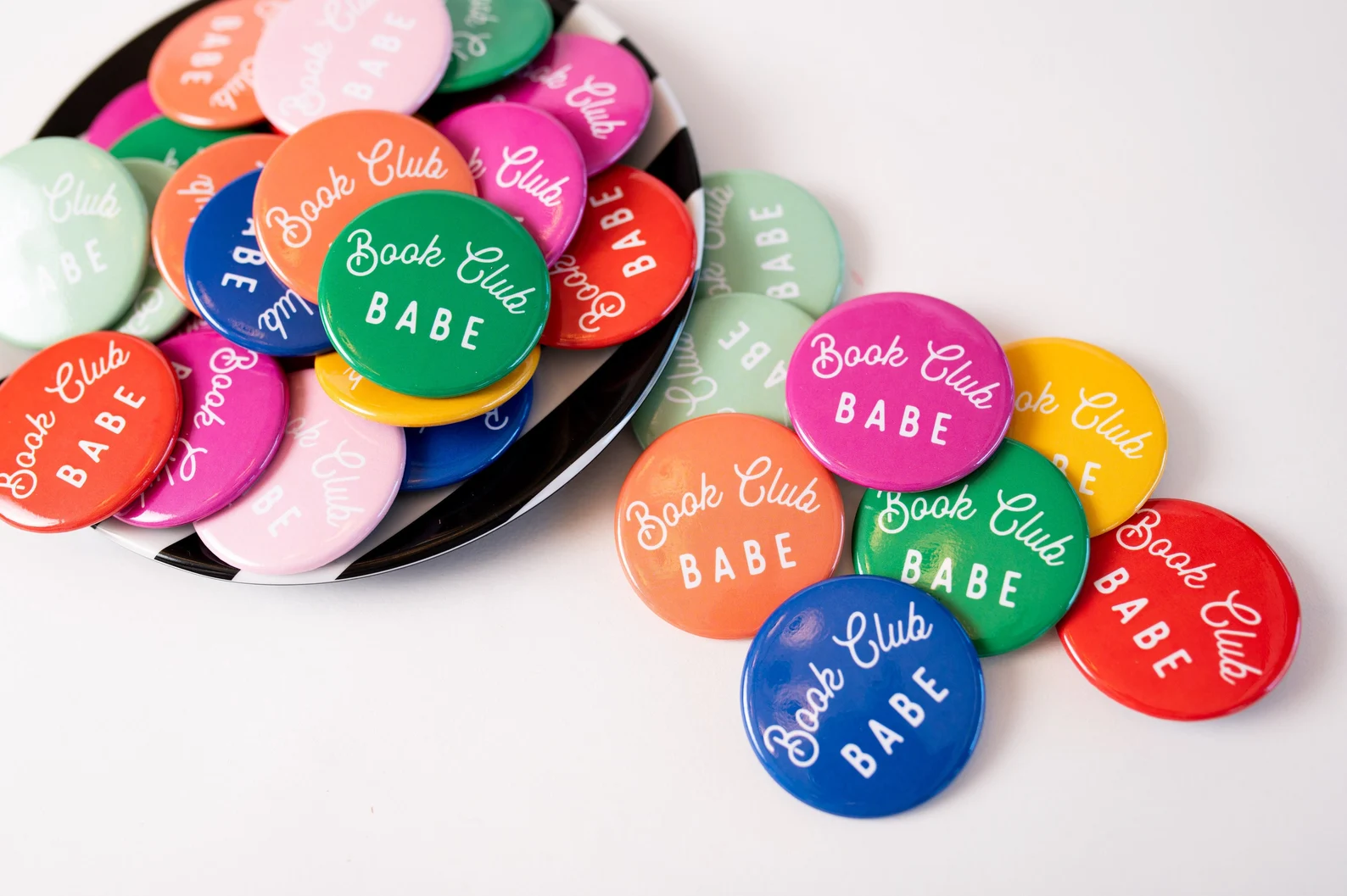 A stack of small colorful round pins that read "book club babe" in cursive script