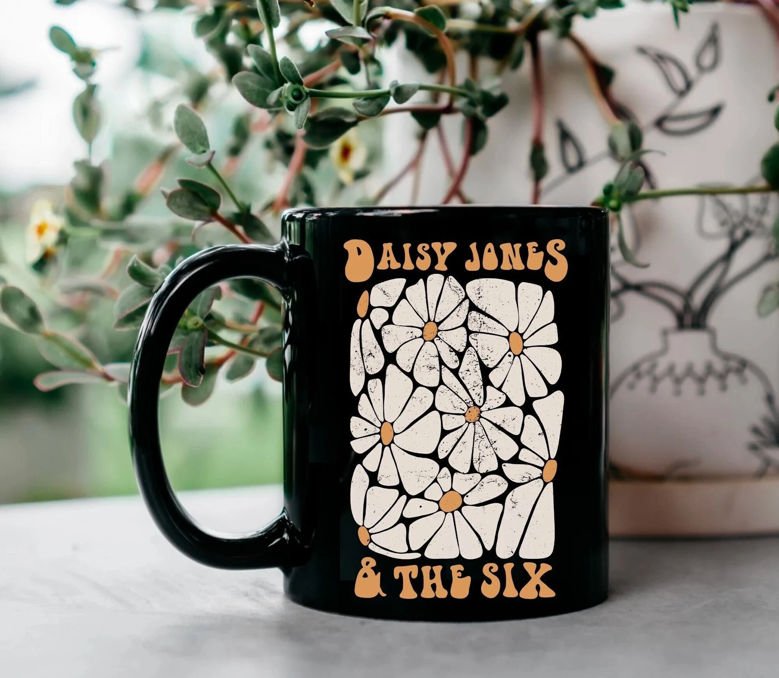 a black mug with white and yellow daisies that reads "Daisy Jones and the Six"