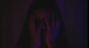 Asian woman in a darkly lit room with her hands over her face