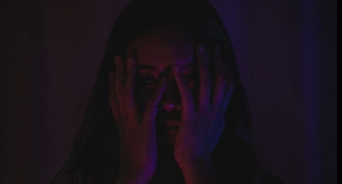 Asian woman in a darkly lit room with her hands over her face