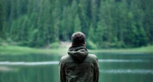 the back of a man wearing a hooded jacket in front of a body of water surrounded by trees