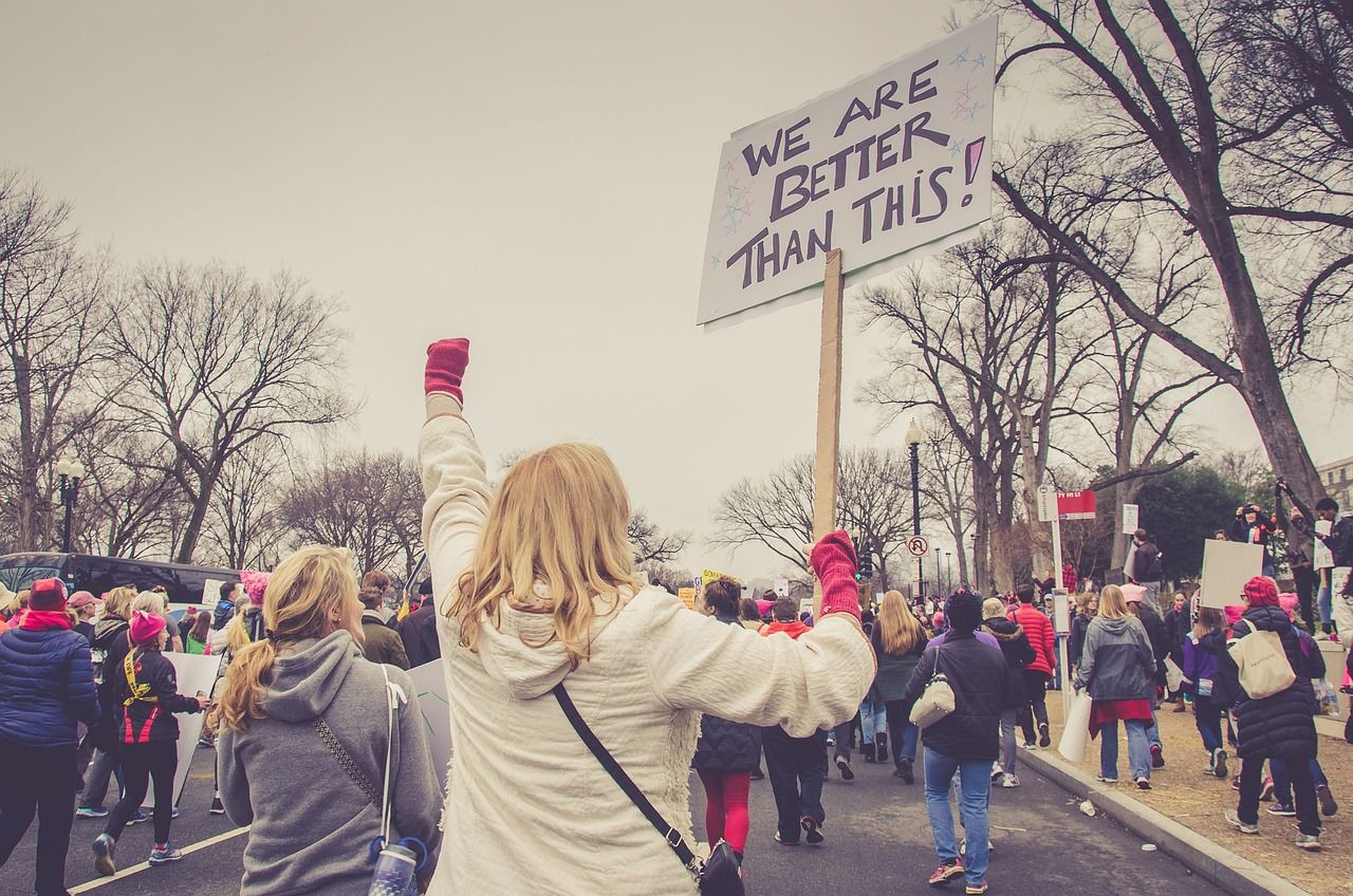 A group of protesters, photographed from behind, walk along a tree-lined road. In the centre of the shot is a person with blonde hair, wearing a white hoodie and pink gloves. They are carrying a placard that reads 'We are better than this!'