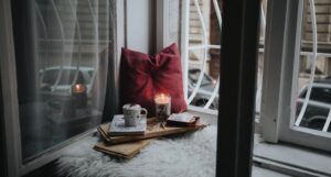reading nook by the window with a mug, a red pillow, a lit candle, and a fuzzy seating pillow