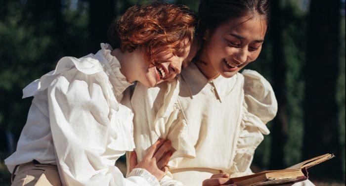 two women- one white and one Asian- laughing and reading together in off white clothes