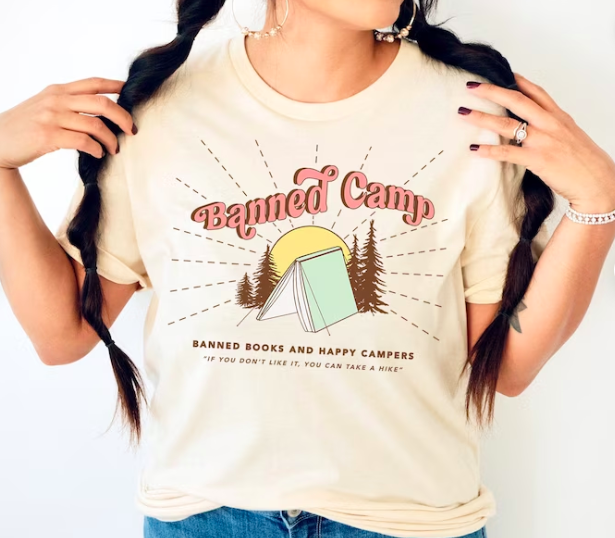 The torso of someone modeling a light orange t-shirt that says "Banned Camp: Banned Books and happy campers, if you don't like it, you can take a hike" and a graphic of a book standing spine-up like a tent surrounded by trees