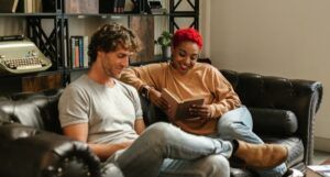 Black woman with short red hair and a white man sit on a couch reading