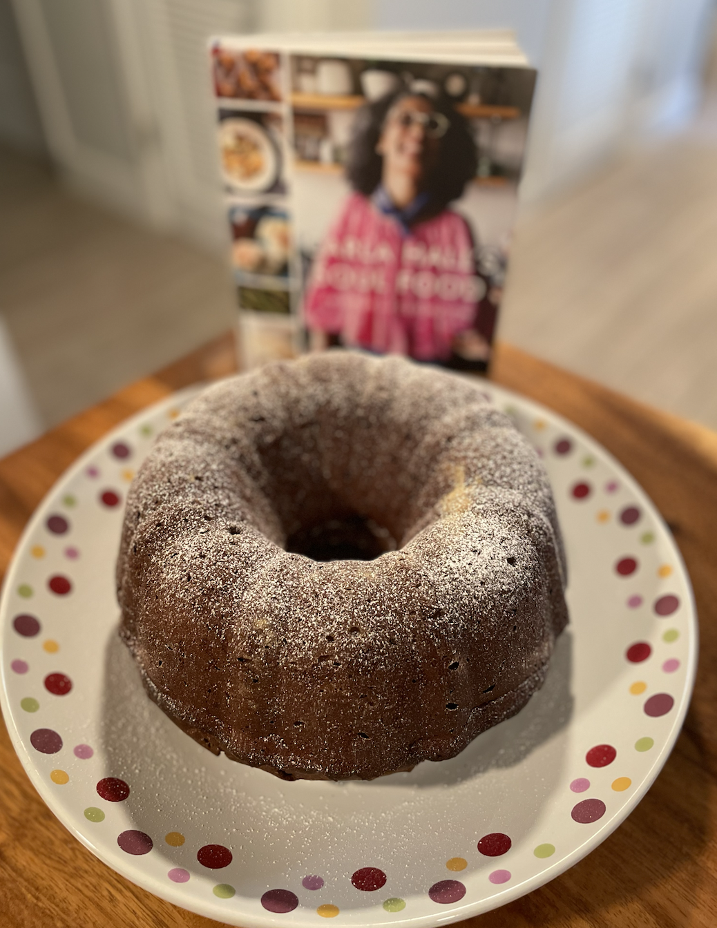 Photo of a chocolate bundt cake dusted with powdered sugar on a dotted serving platter on a wooden table with the cookbook Carla Hall's Soul Food