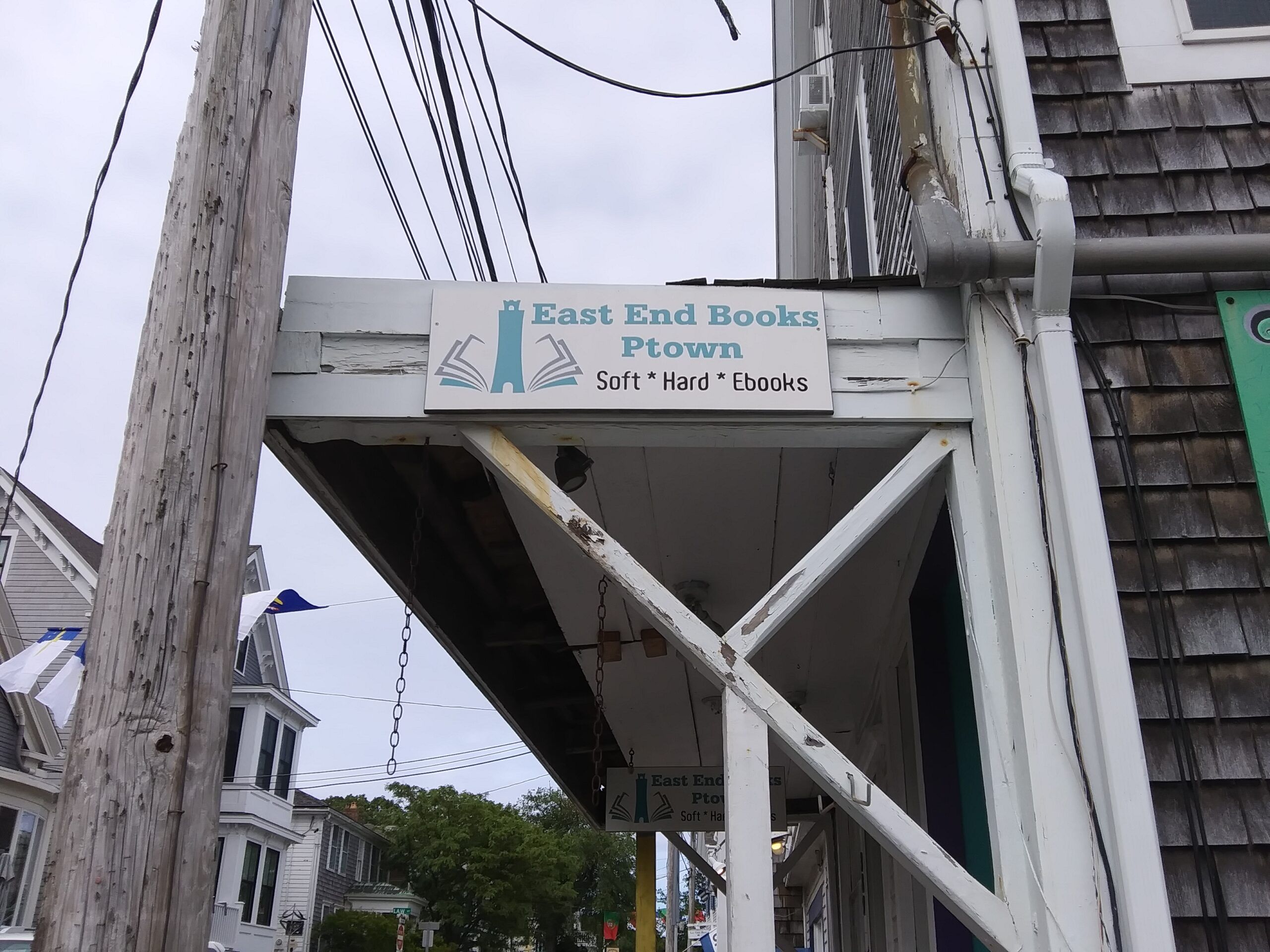 A sign affixed to the side of a horizontal awning. The sign says East End Books Ptown" in pale blue letters, along with the logo of a lighthouse flanked by book pages