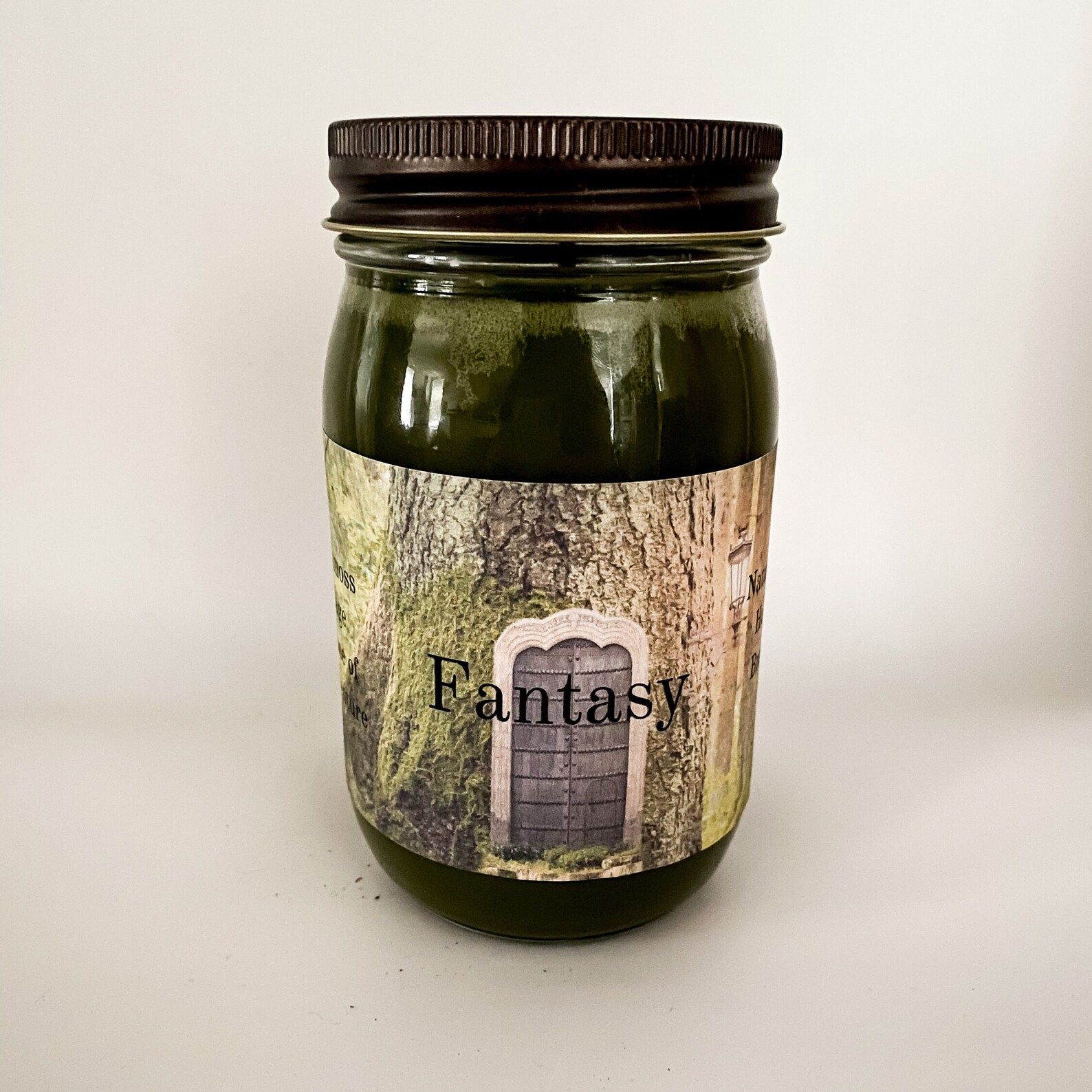 Green candle in a clear jar with a black lid. Label with Fantasy written over a tree with a door.